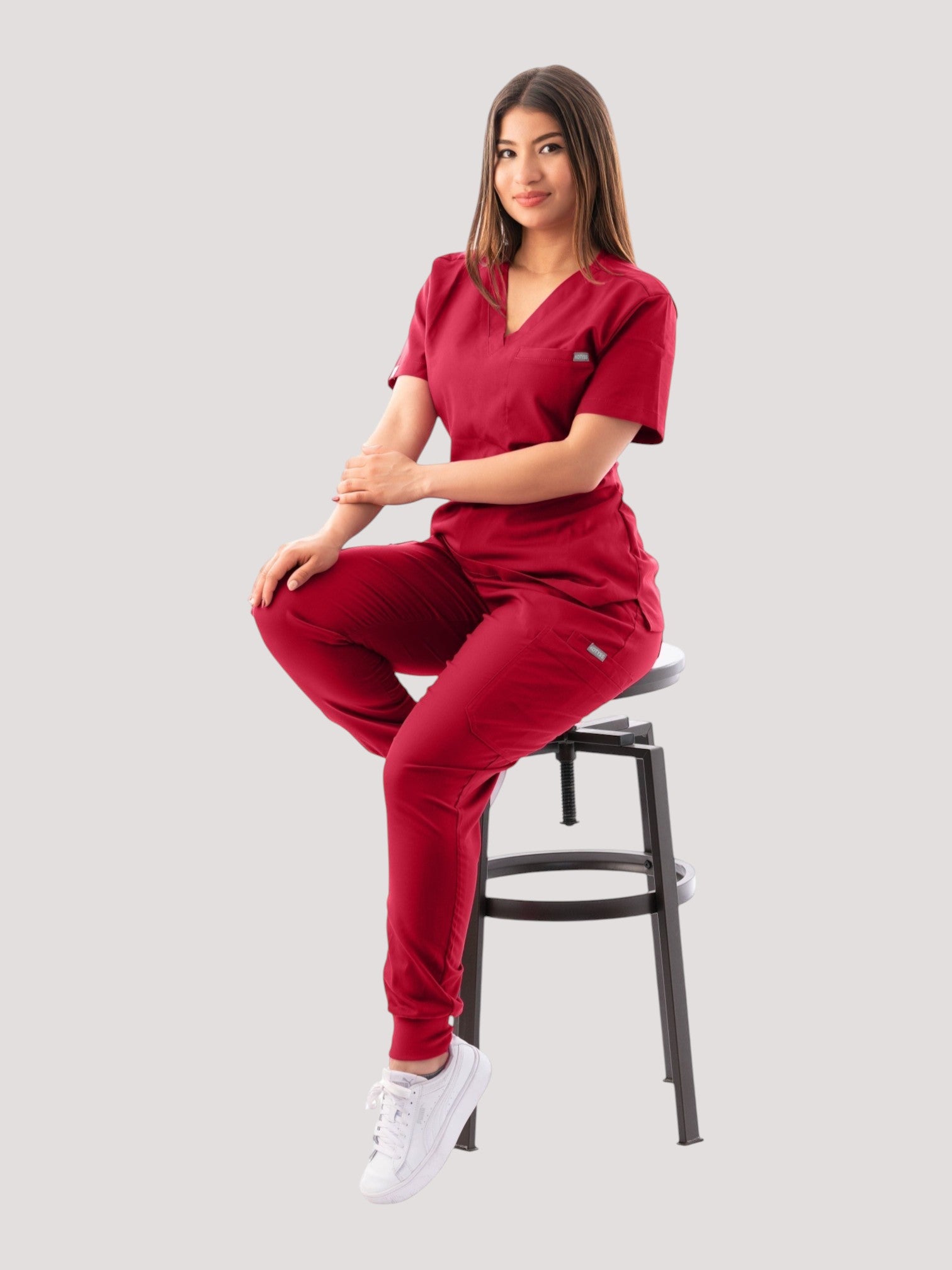 A woman wearing a deep red Kotyss V-neck scrub top and matching jogger pants while sitting on a stool.