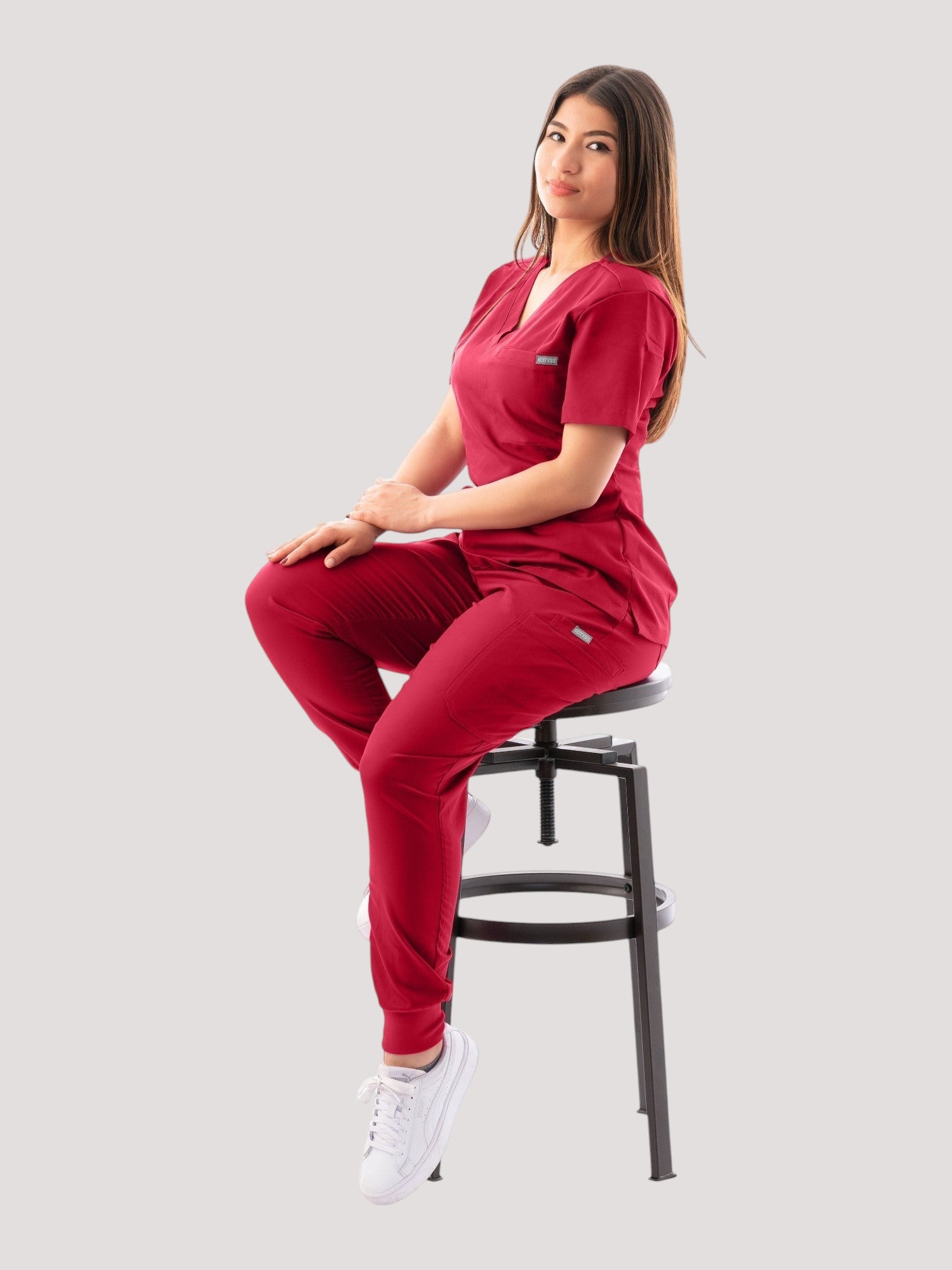 A woman wearing a deep red Kotyss V-neck scrub top and matching jogger pants, sitting on a stool.