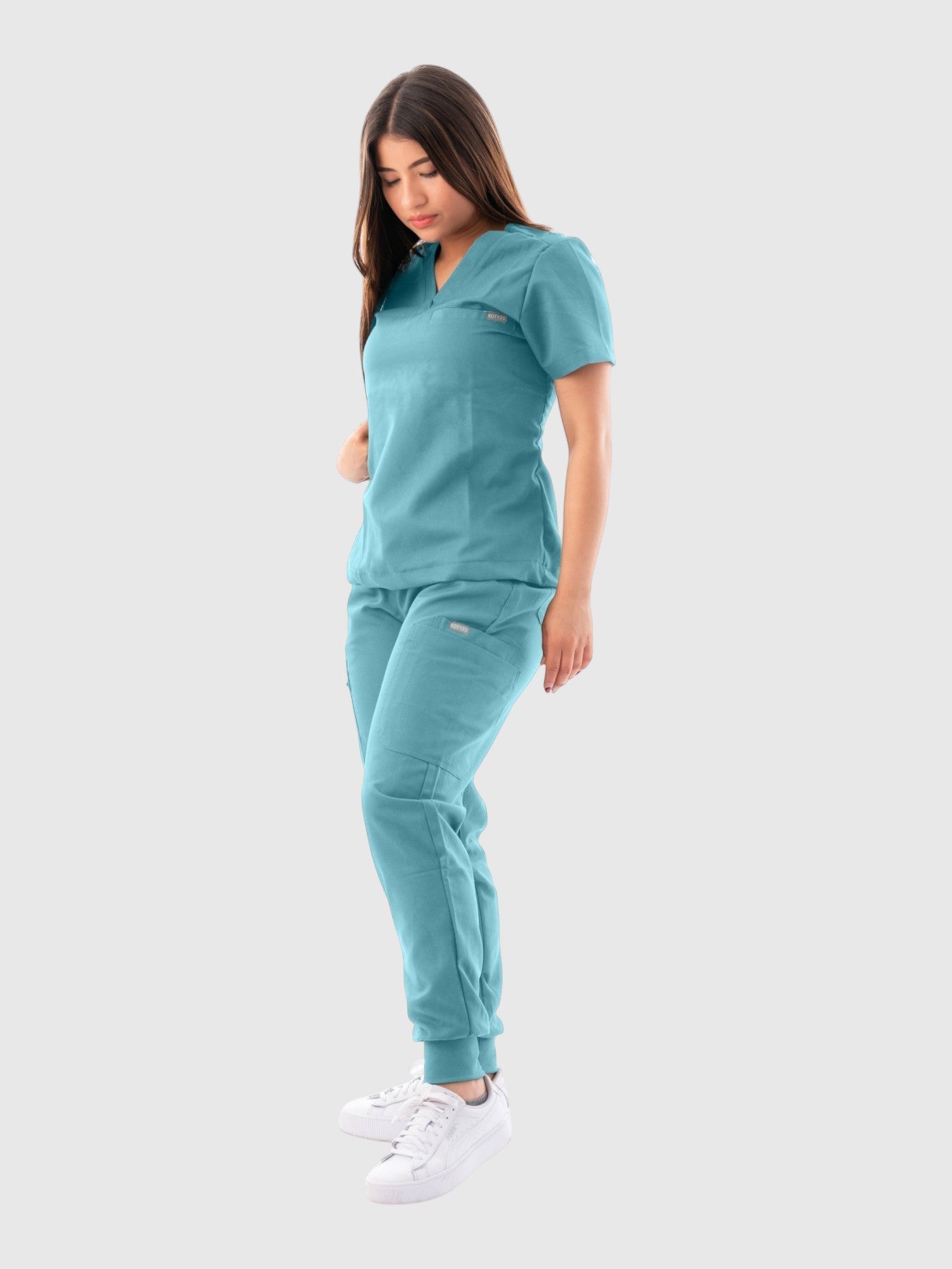 Model wearing a teal Kotyss V-neck scrub top and matching jogger pants with multiple pockets for medical professionals