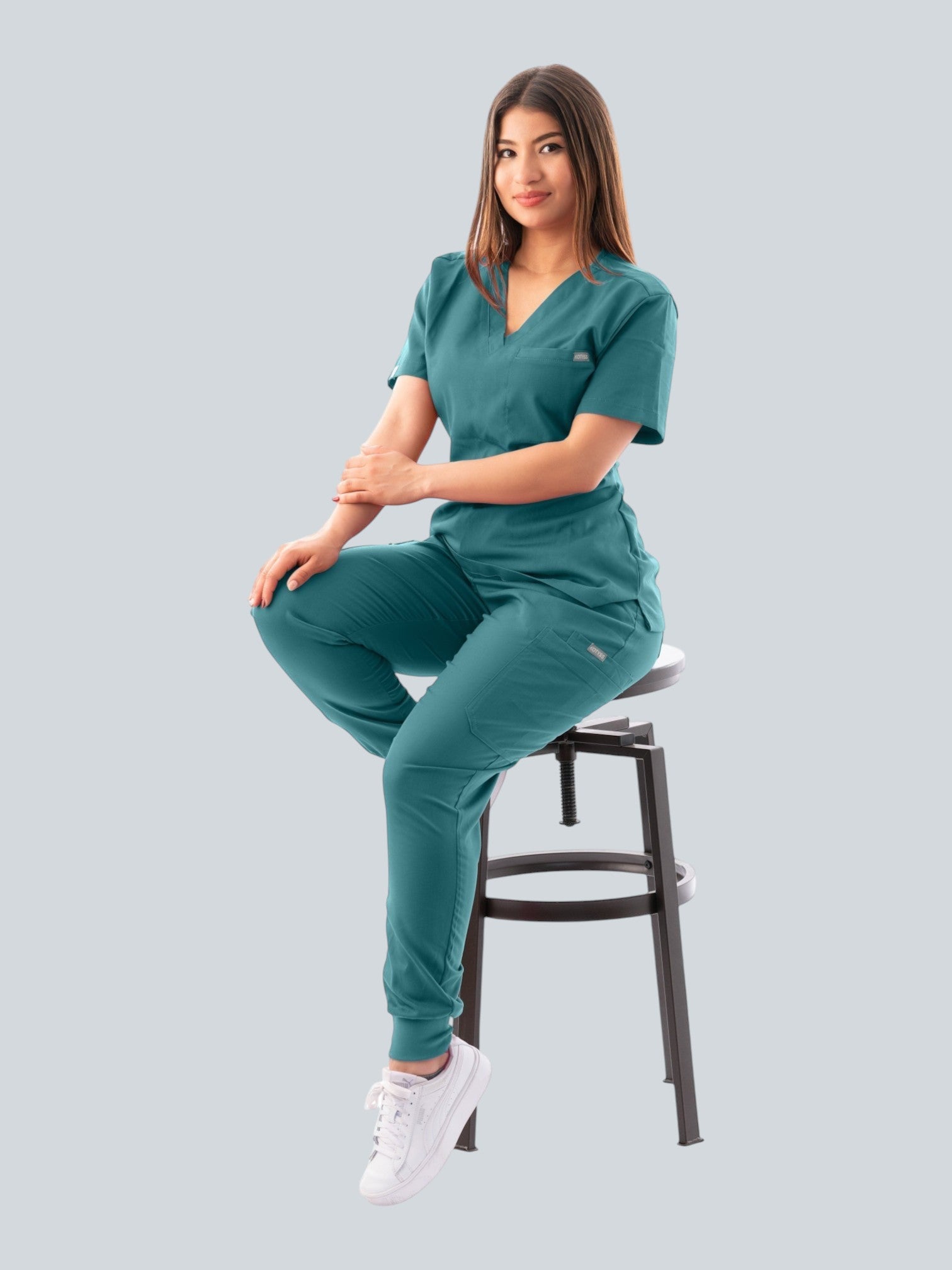 Model wearing a teal Kotyss V-neck scrub top and jogger pants set, featuring multiple pockets and 4-way stretch fabric.
