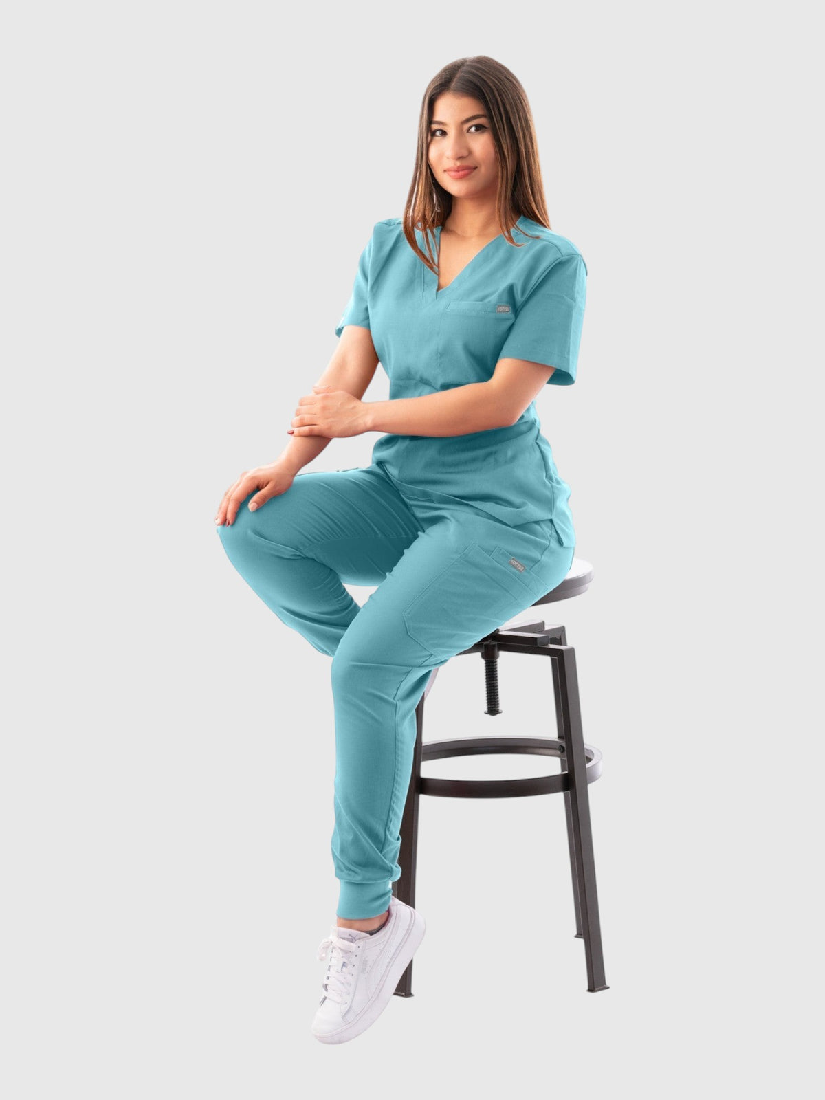 A woman wearing a teal Kotyss V-neck scrub top and matching jogger pants, sitting on a stool in a studio setting.