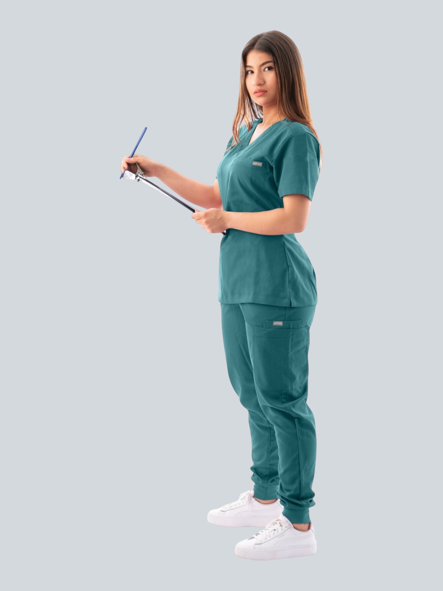 A woman wearing a teal Kotyss V-neck scrub top and jogger pants, holding a clipboard against a grey background.