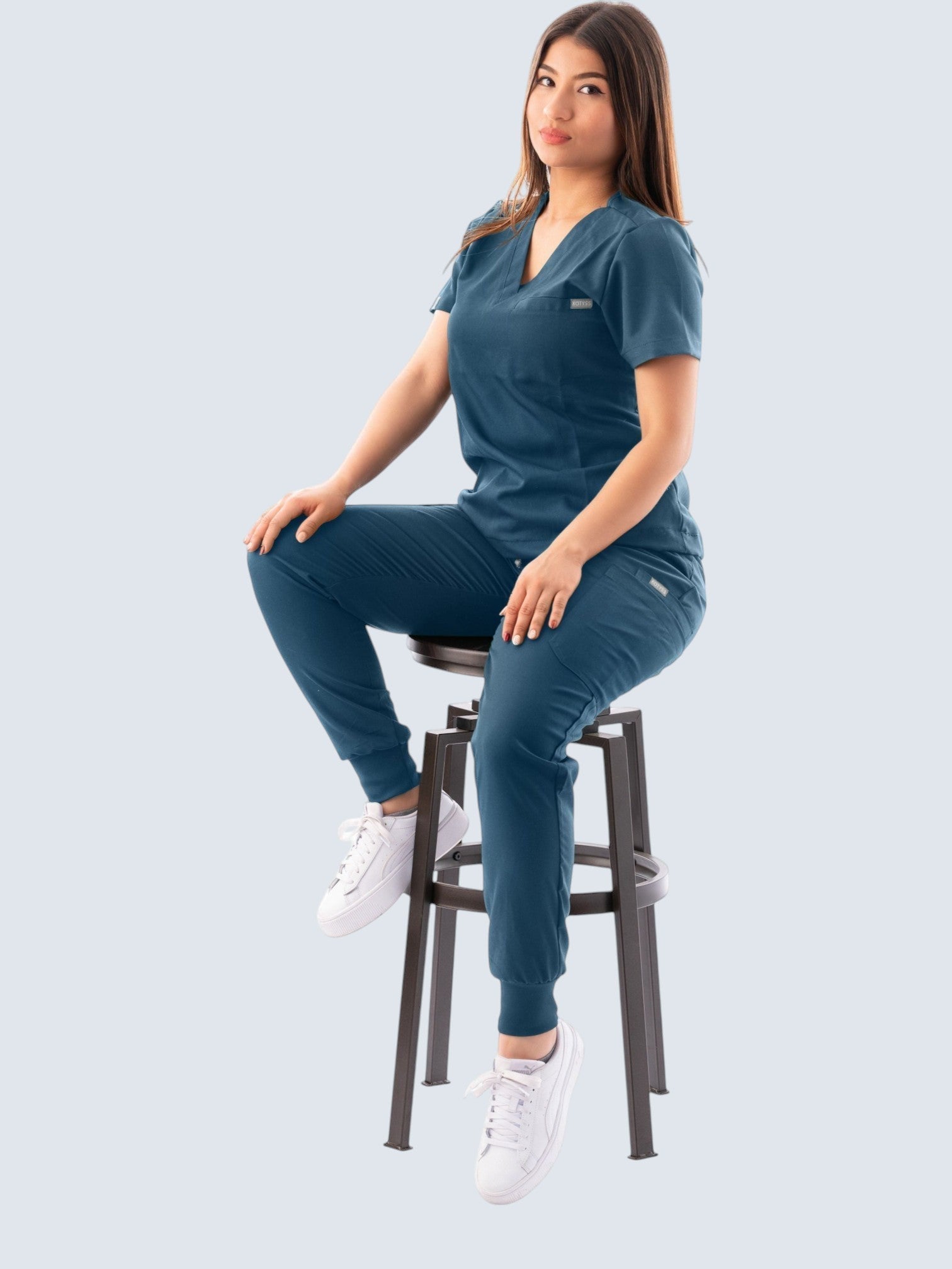 Kotyss women's teal V-neck scrub top and jogger pants set worn by a model sitting on a stool.