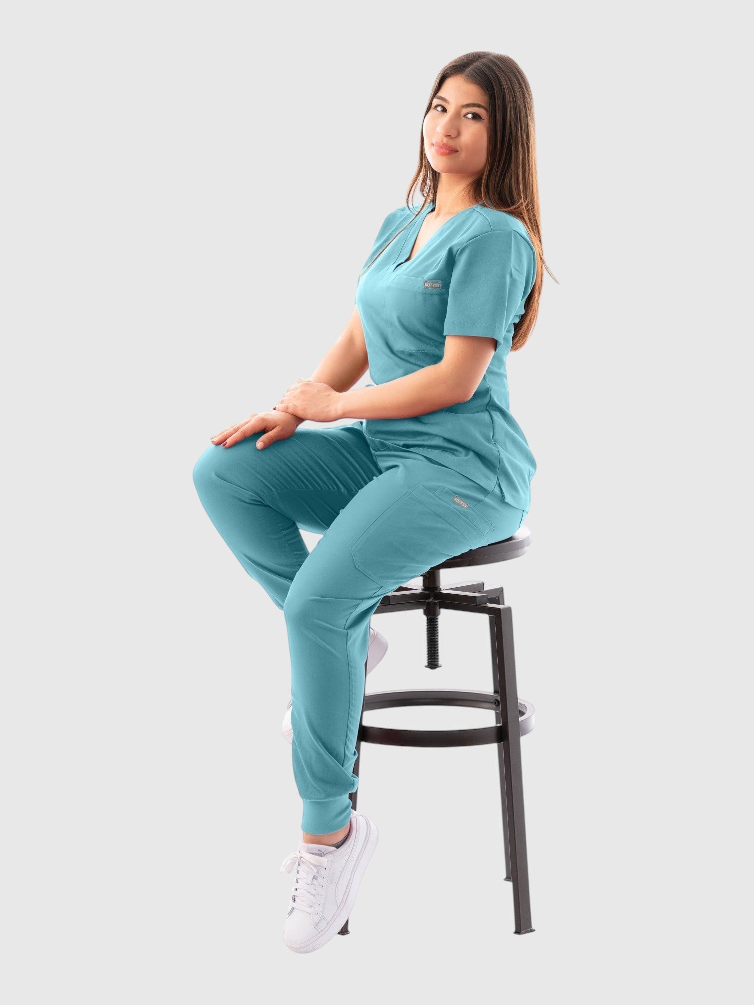 Kotyss women's teal V-neck scrub top and jogger pants set with 4-way stretch fabric worn by a model on a stool.