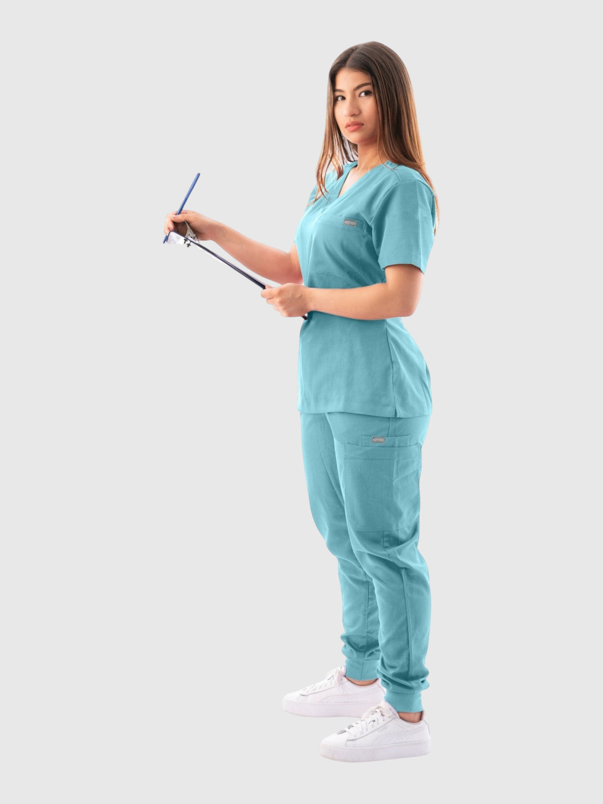 A woman wearing a teal Kotyss V-neck scrub top and jogger pants set, holding a clipboard in a studio setting.