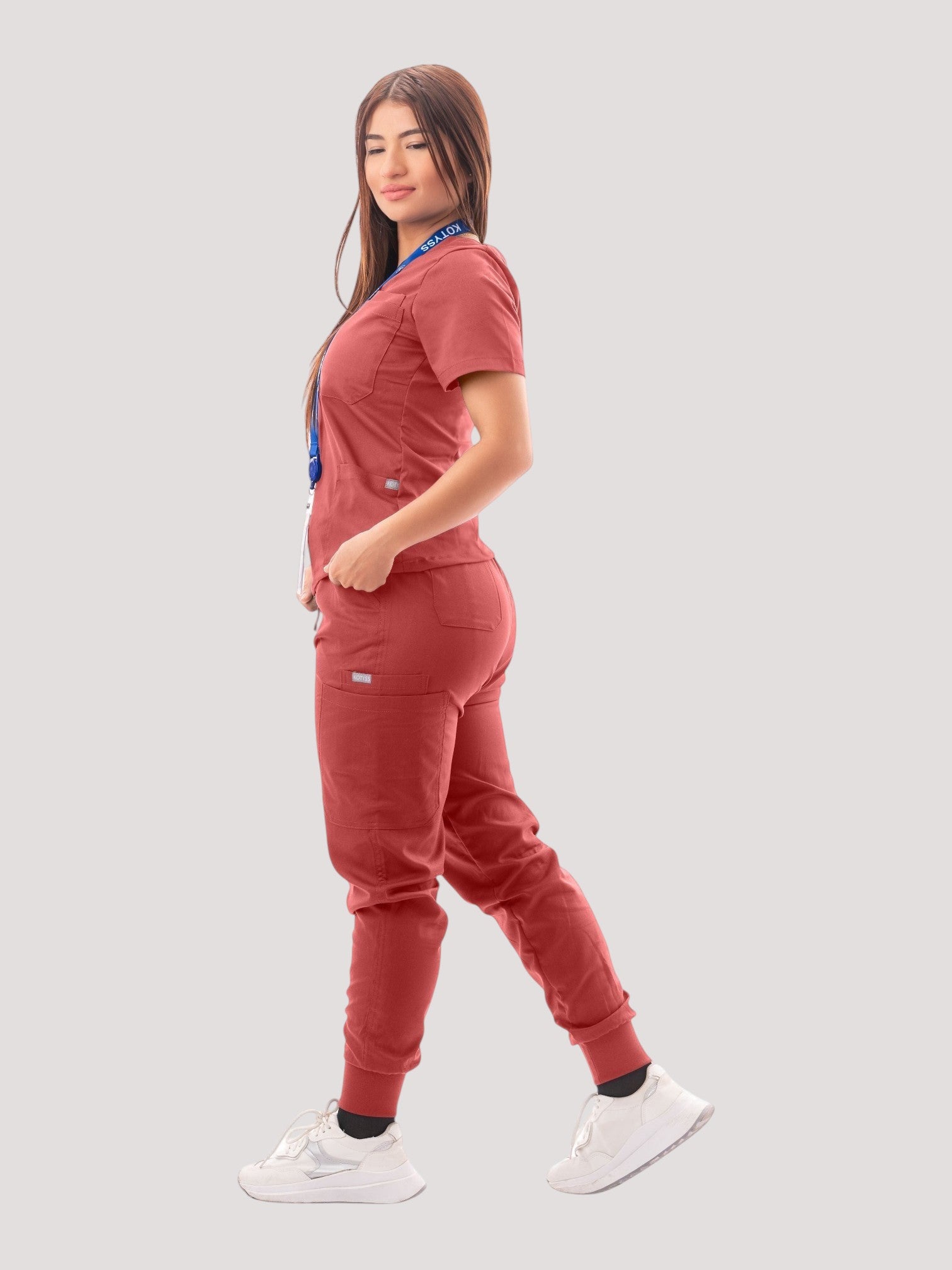 A woman wearing a terracotta Kotyss V-neck scrub top and multi-pocket jogger pants set with white sneakers.