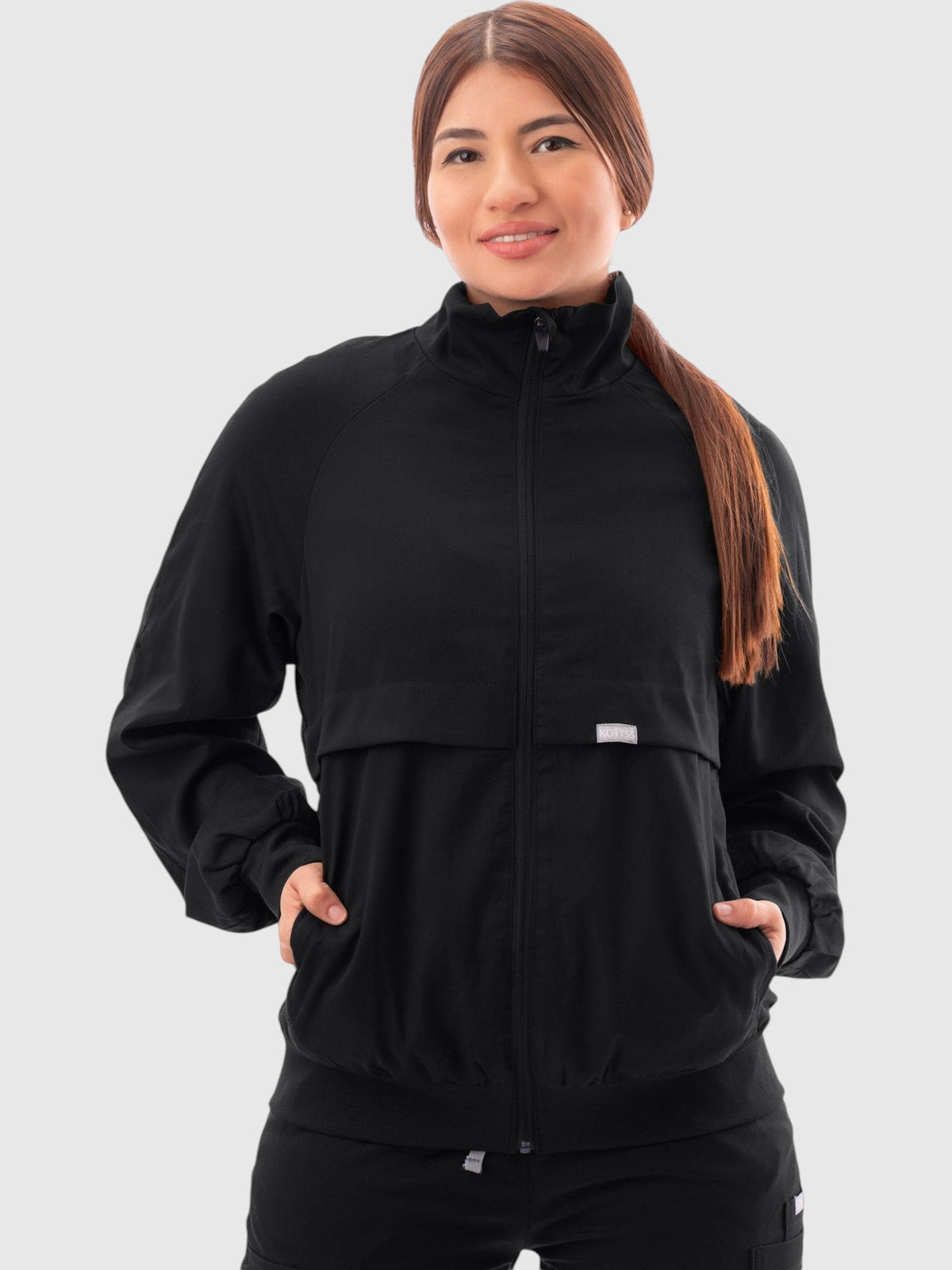 Kotyss women's black scrub jacket with a high windbreaker neck, zip front, and ribbed cuffs on a professional model.